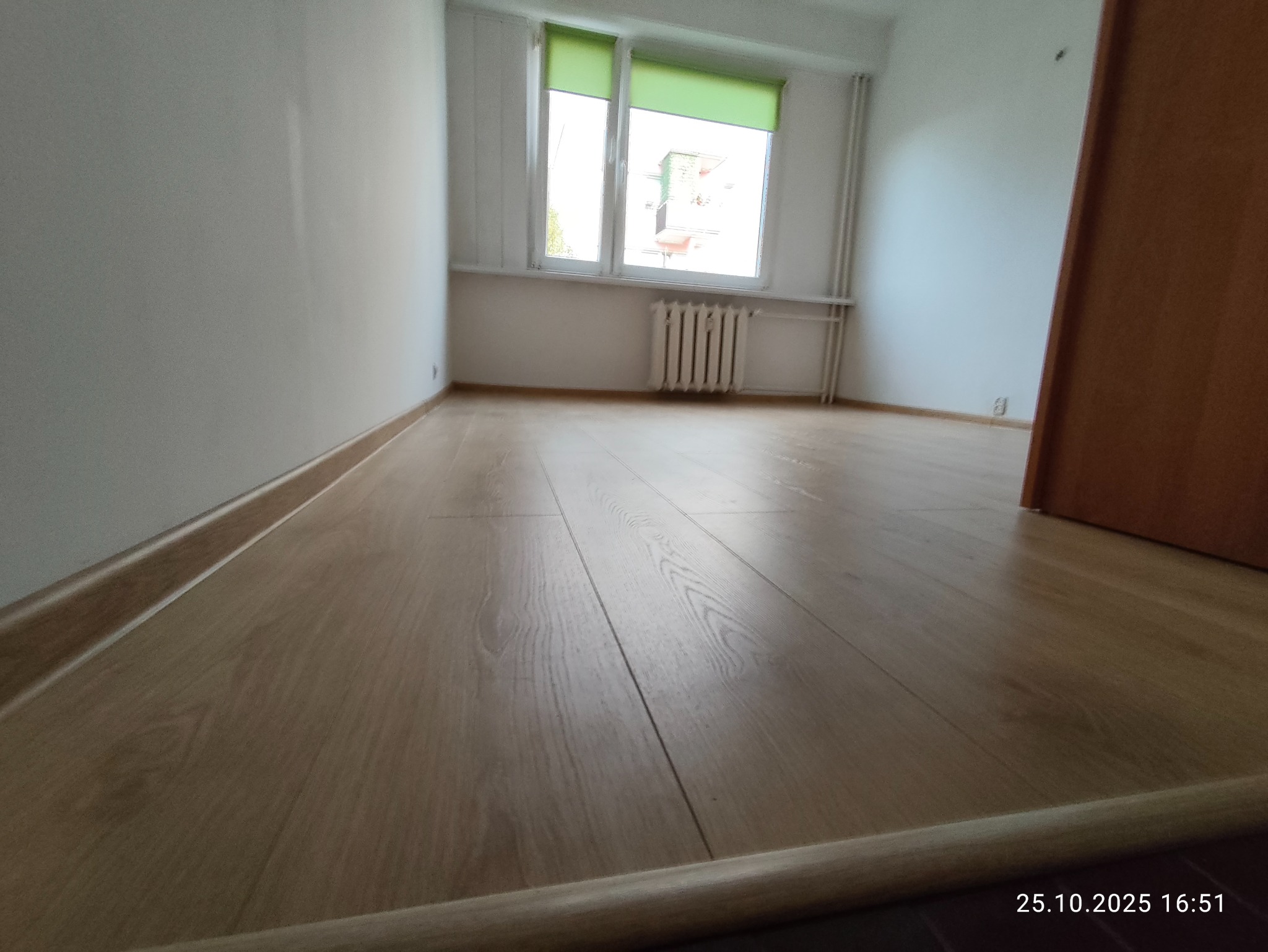 Jasne panele podłogowe w pokoju po remoncie w Nysie. Widok na okno z zieloną roletą i grzejnik. Świeżo wykończone wnętrze z listwami przypodłogowymi.