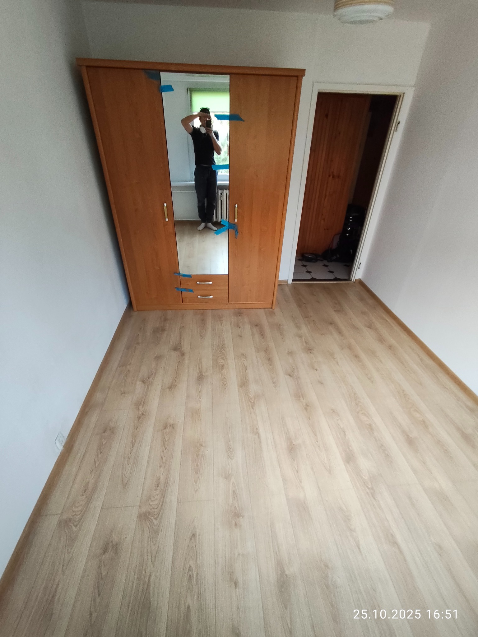 Jasna podłoga z paneli, drewniana szafa z lustrem zabezpieczona taśmą, białe ściany i widok na sąsiednie pomieszczenie. Ujęcie szerokokątne.