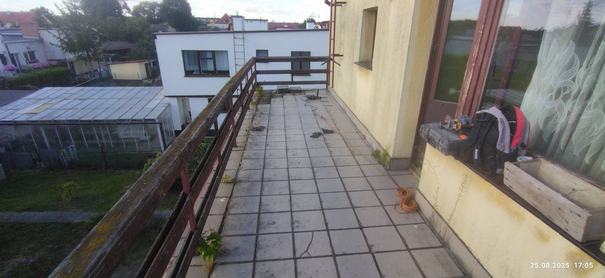 Balkon z szarą, kwadratową terakotą w trakcie remontu. Widoczne narzędzia, kabel i torba. Drewniana balustrada i budynki w tle. Ujęcie z góry.