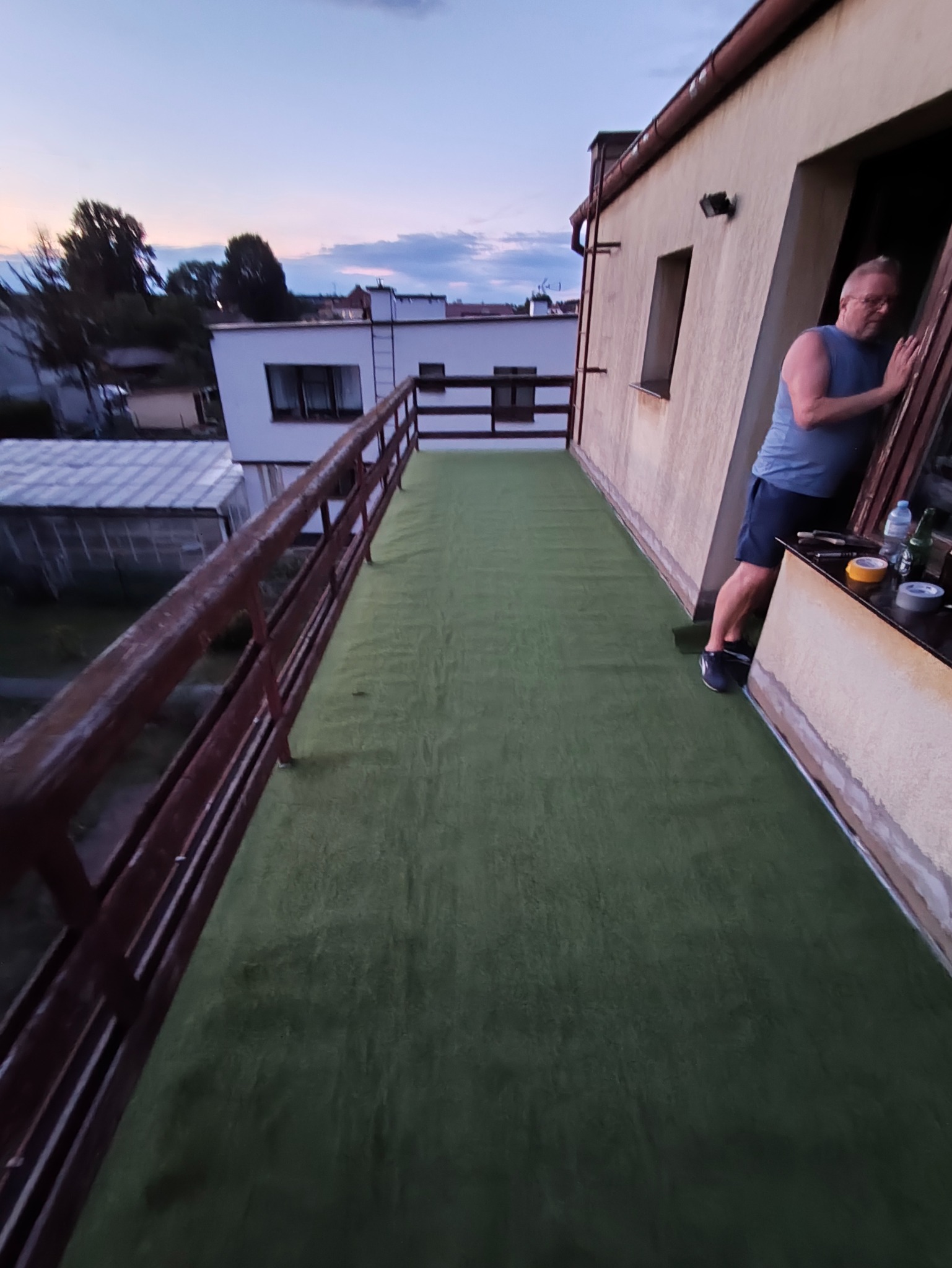 Zielony dywan na balkonie z drewnianą balustradą. Mężczyzna w tle opiera się o framugę okna. Widok na budynki i niebo o zmierzchu.