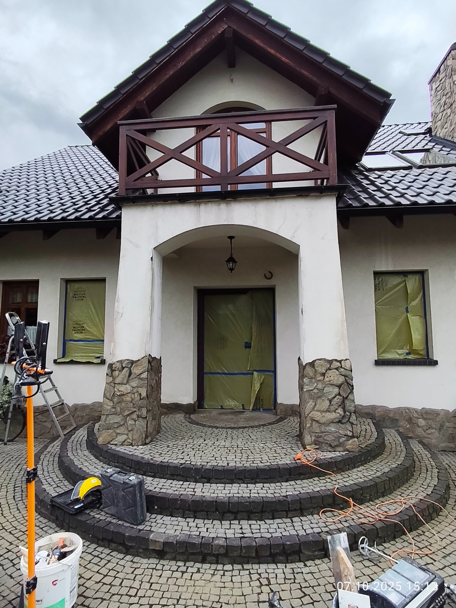 Dom jednorodzinny w trakcie przygotowań do malowania elewacji, okna i drzwi zabezpieczone folią, widoczne narzędzia i materiały malarskie.