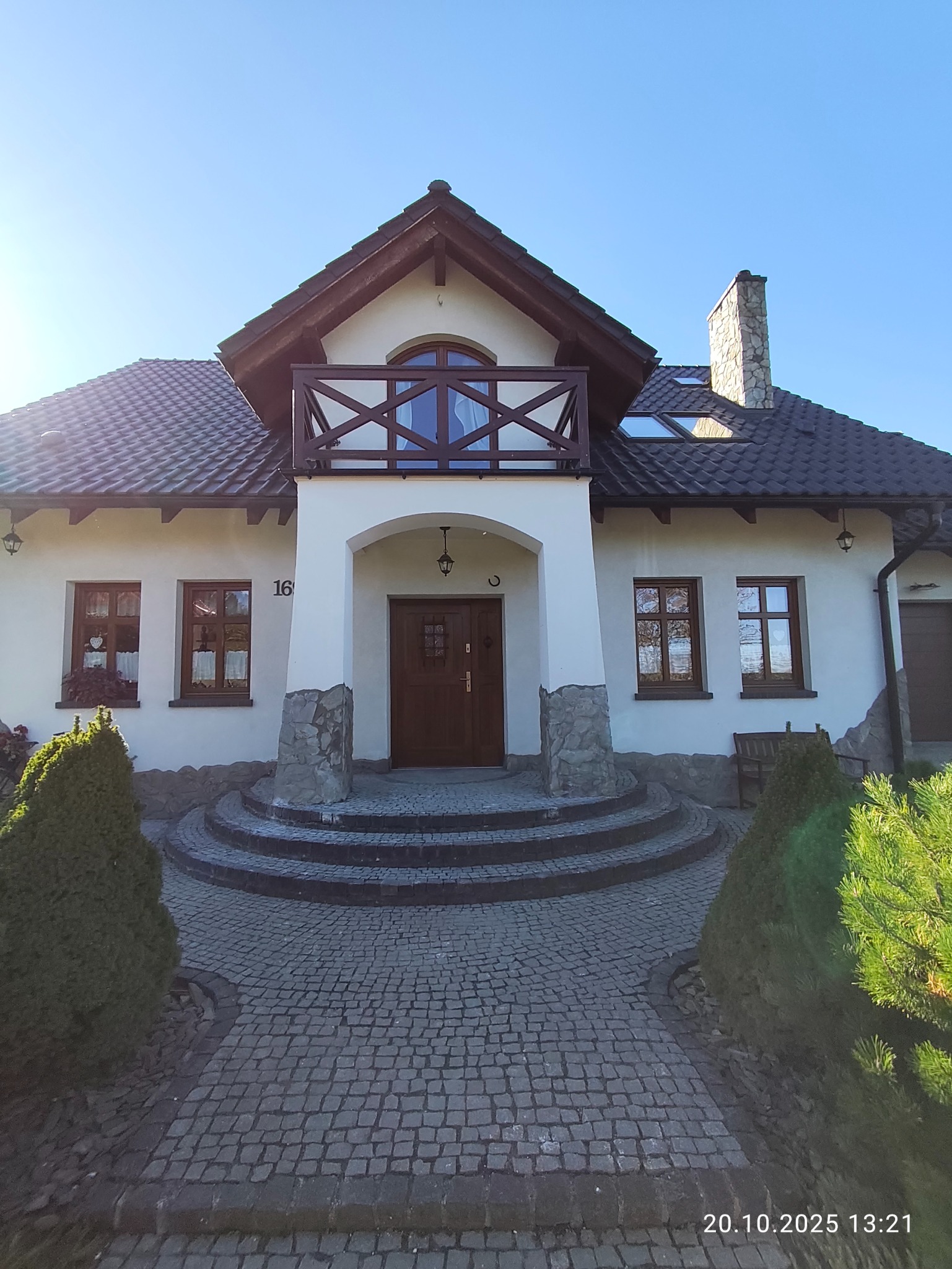 Elewacja domu jednorodzinnego z balkonem i kamiennym wykończeniem, widok z poziomu chodnika. Uwagę przykuwa drewniana stolarka okienna i drzwiowa.