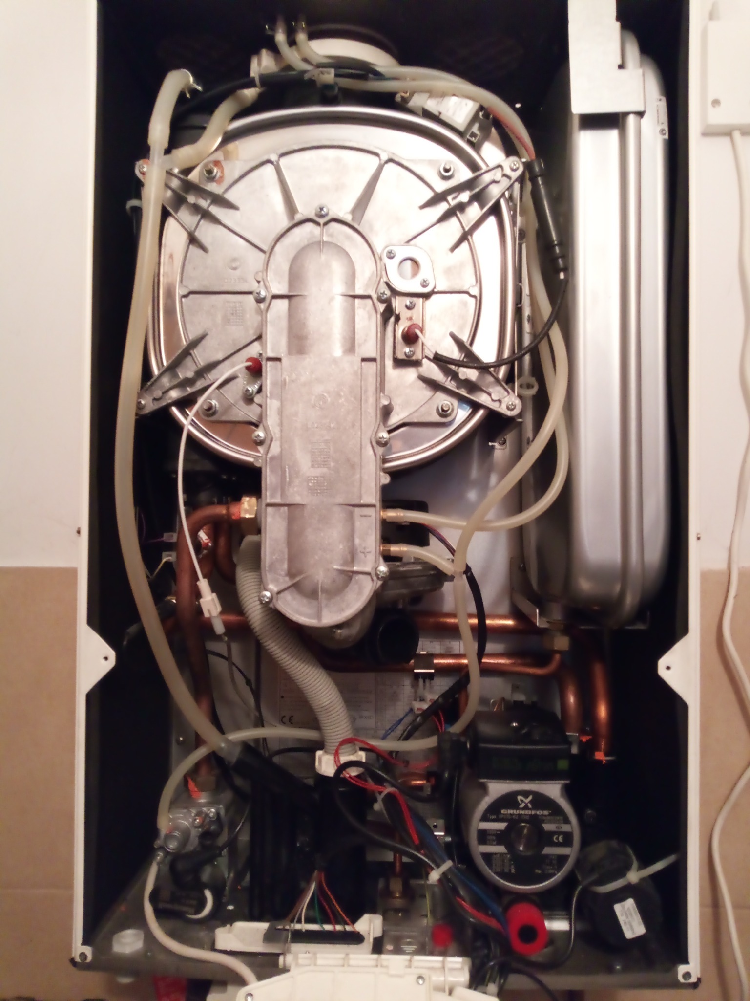 Otwarty piec gazowy z widocznymi elementami wewnętrznymi: rurami miedzianymi, przewodami, pompą obiegową Grundfos i srebrnym zbiornikiem wyrównawczym. Widok z przodu.