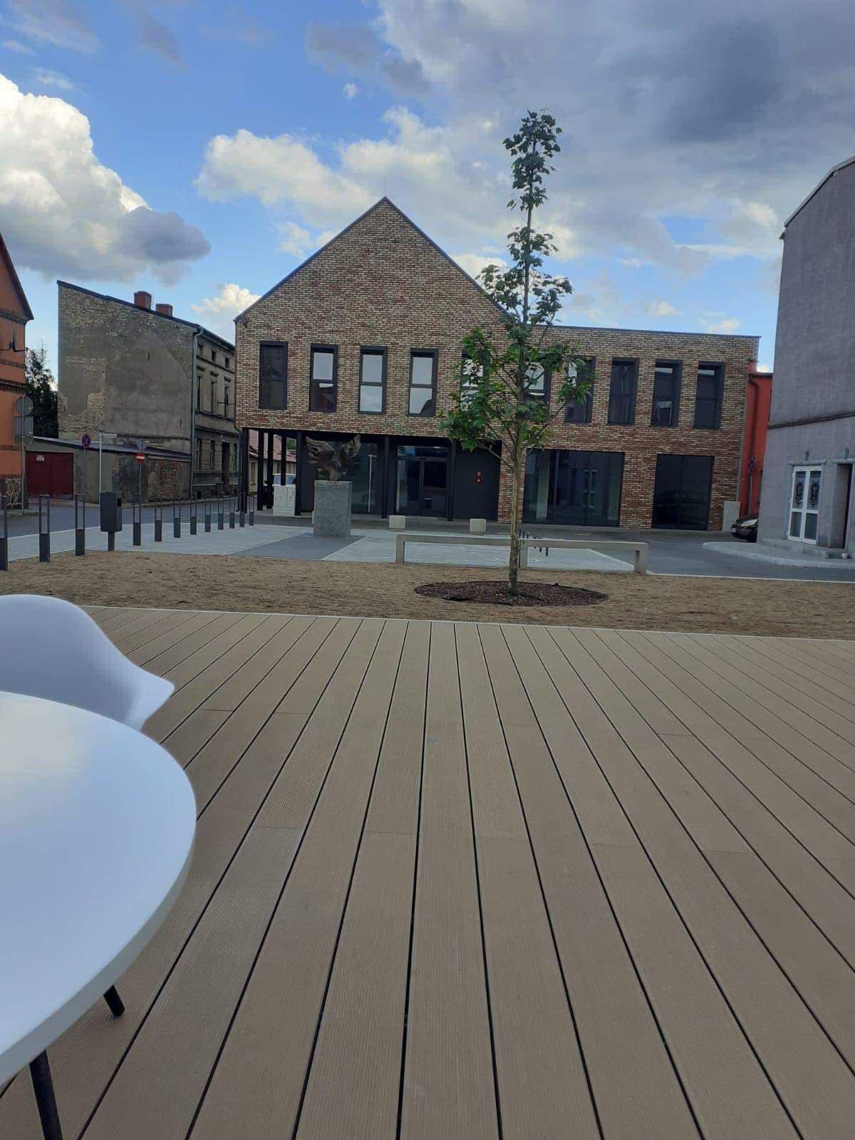 Nowoczesny budynek z cegły z ciemnymi oknami, widok z drewnianego tarasu z białymi krzesłami. Uwagę przykuwa rzeźba orła przed budynkiem i młode drzewo.