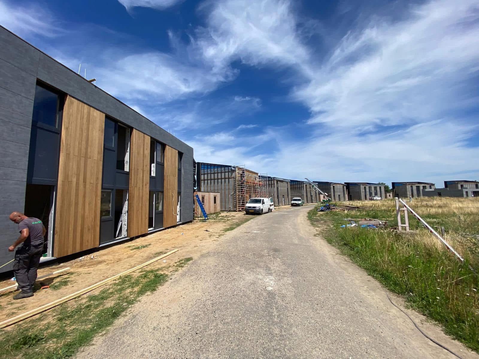 Nowoczesne budynki w trakcie montażu elewacji z drewnianymi panelami, widoczne rusztowania i pojazdy budowlane, błękitne niebo z chmurami w tle.