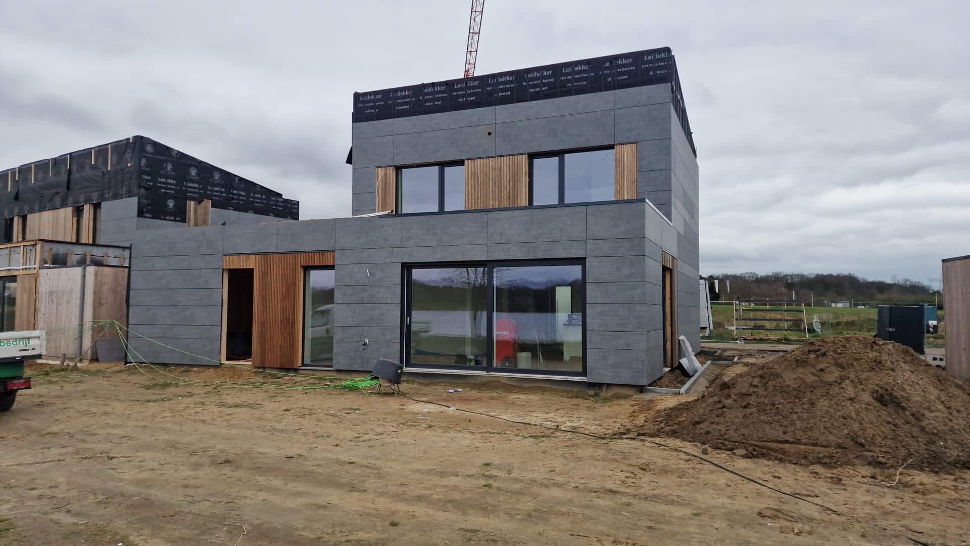 Nowoczesny dom w trakcie wykończenia elewacji z szarych paneli i drewnianych akcentów. Widoczne duże przesuwne okno i elementy konstrukcyjne. Na pierwszym planie ziemia i sprzęt.