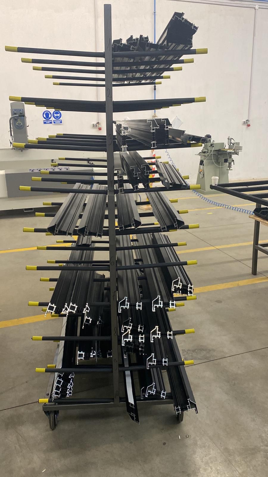 Wózek z czarnymi profilami aluminiowymi z żółtymi zabezpieczeniami, przygotowanymi do dalszej obróbki w hali produkcyjnej. Widoczna maszyna w tle.