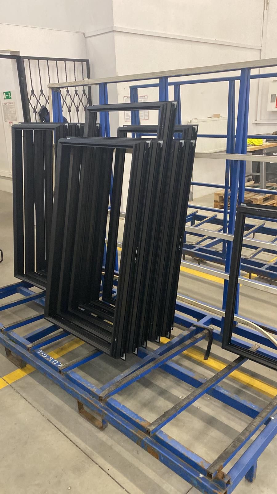 Stos czarnych ram okiennych na niebieskim wózku transportowym w hali produkcyjnej. Widoczne detale konstrukcyjne ram i numer seryjny na wózku.