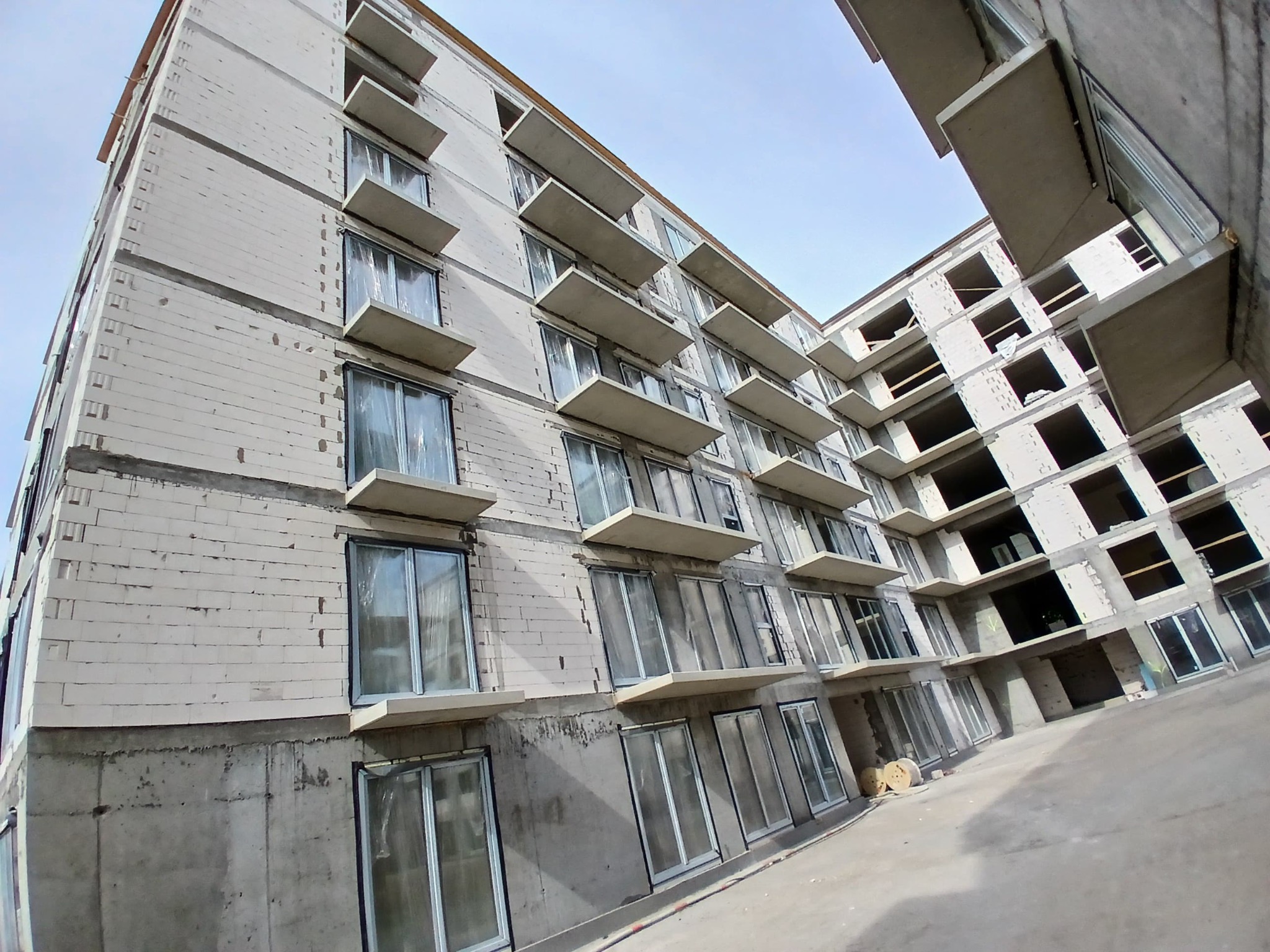 Wielopiętrowy budynek w stanie surowym z zamontowanymi oknami, widok z dołu pod kątem, ukazujący powtarzalność balkonów i okien.