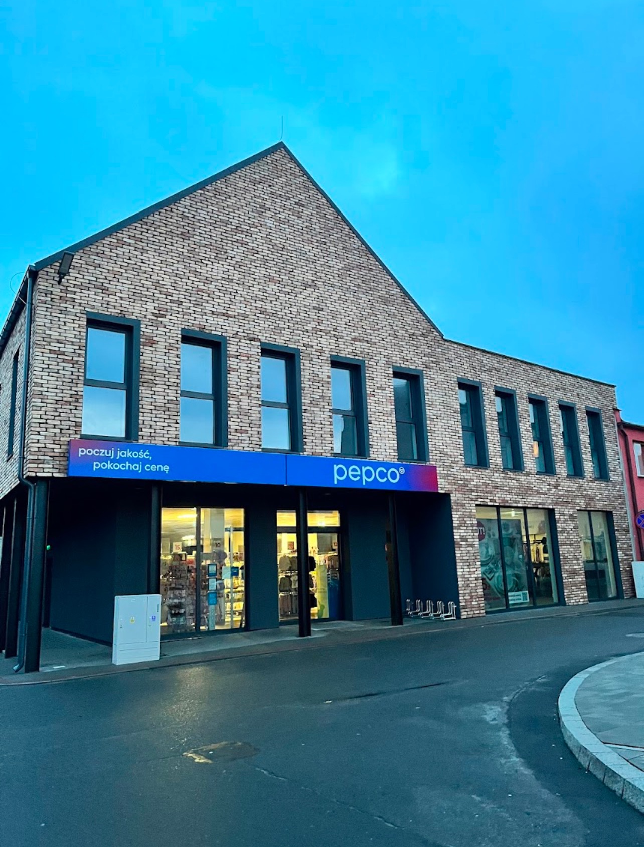 Fasada budynku Pepco z cegły klinkierowej, z czarnymi ramami okiennymi i niebieskim szyldem. Widoczne witryny sklepowe i fragment ulicy.