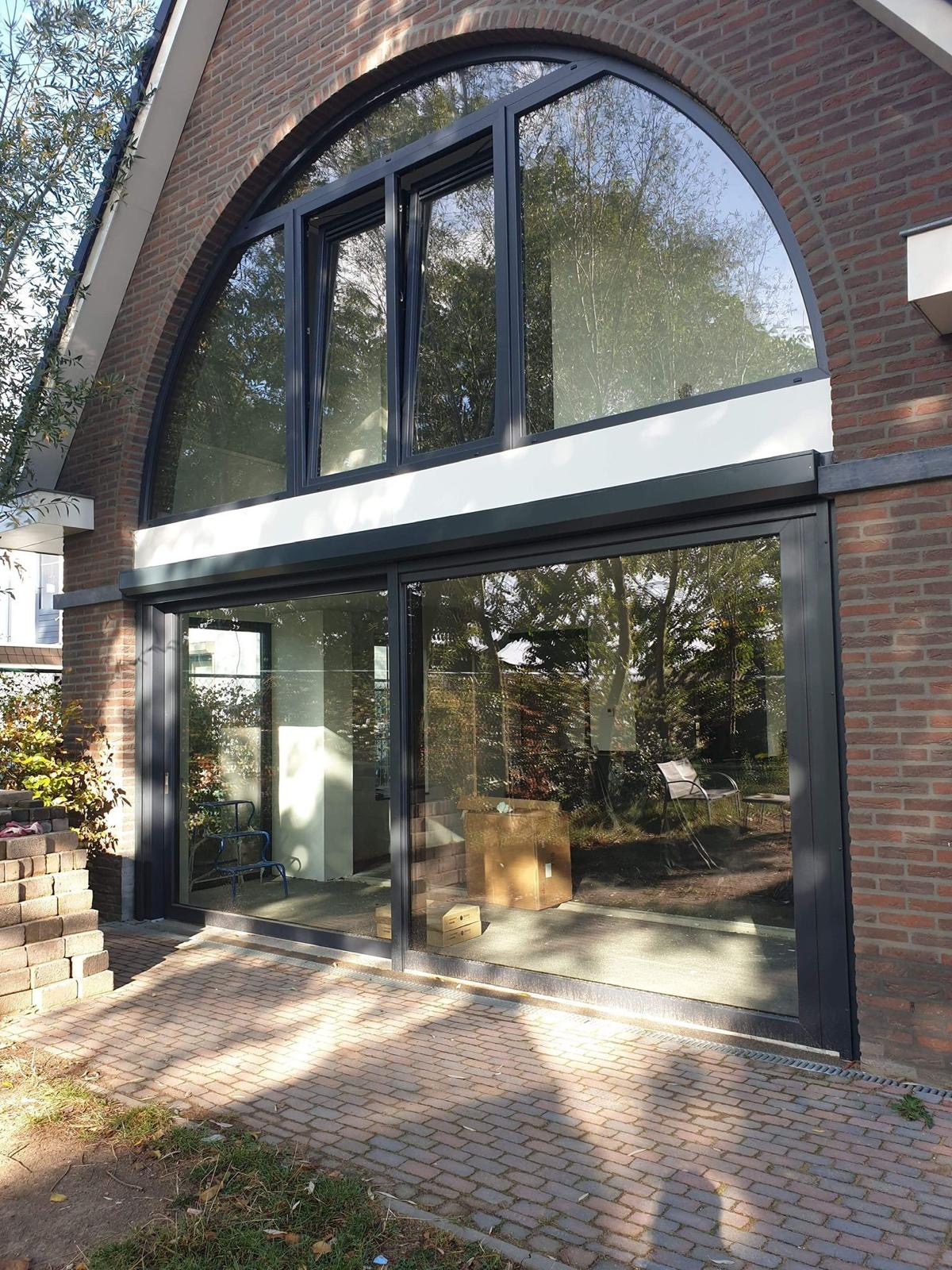 Nowoczesna fasada domu z cegły z dużymi, czarnymi oknami o nietypowym kształcie łuku i przesuwnymi drzwiami, odbijającymi drzewa i niebo.