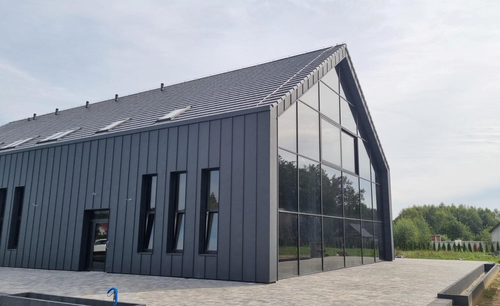 Nowoczesny budynek z grafitową elewacją, dużymi przeszkleniami i skośnym dachem z oknami połaciowymi. Widok z zewnątrz, kostka brukowa na pierwszym planie.