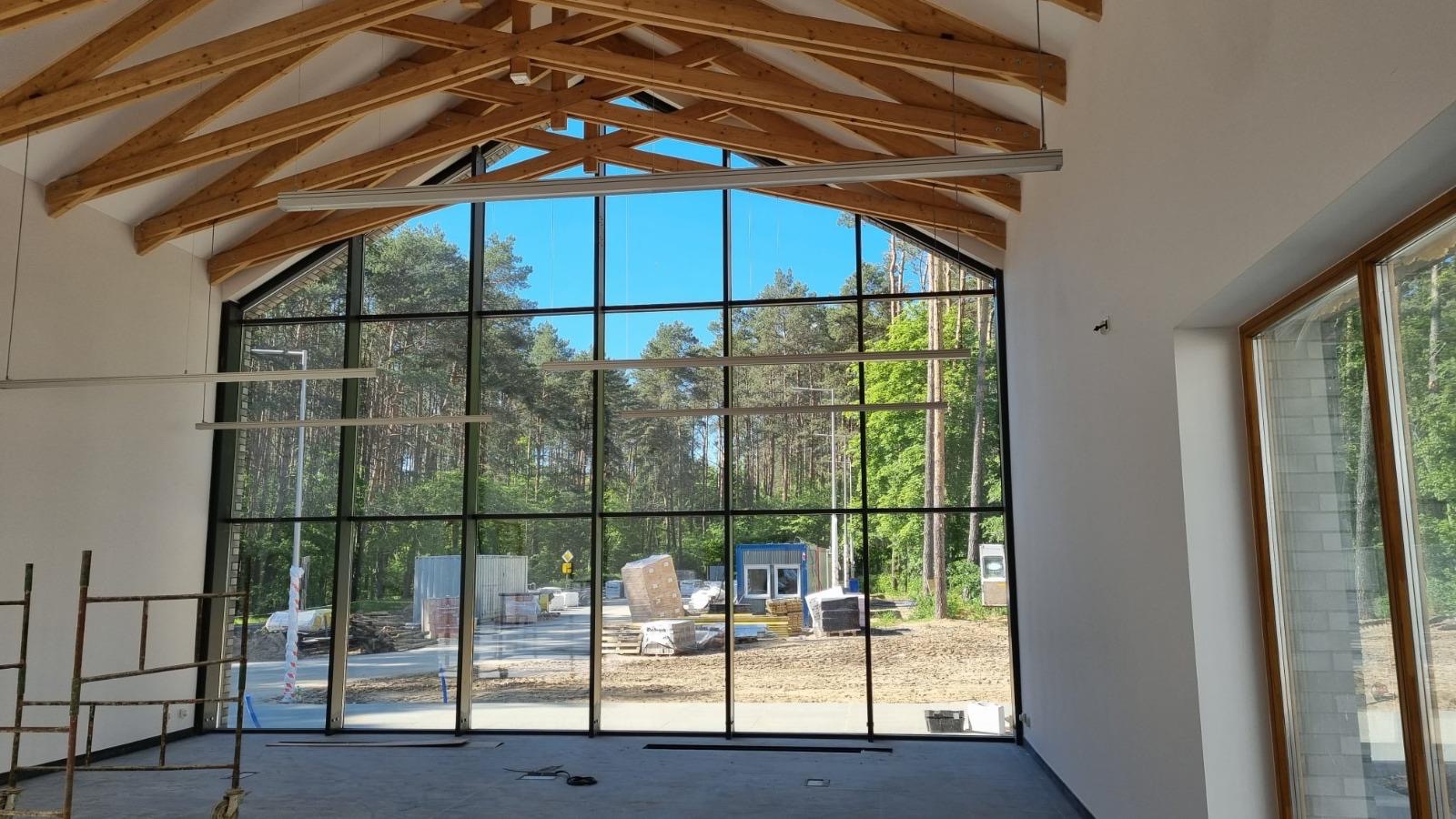 Wysokie okno z czarnymi szprosami w surowym wnętrzu z drewnianą konstrukcją dachu. Widok na las i plac budowy. Częściowo widoczne rusztowanie.