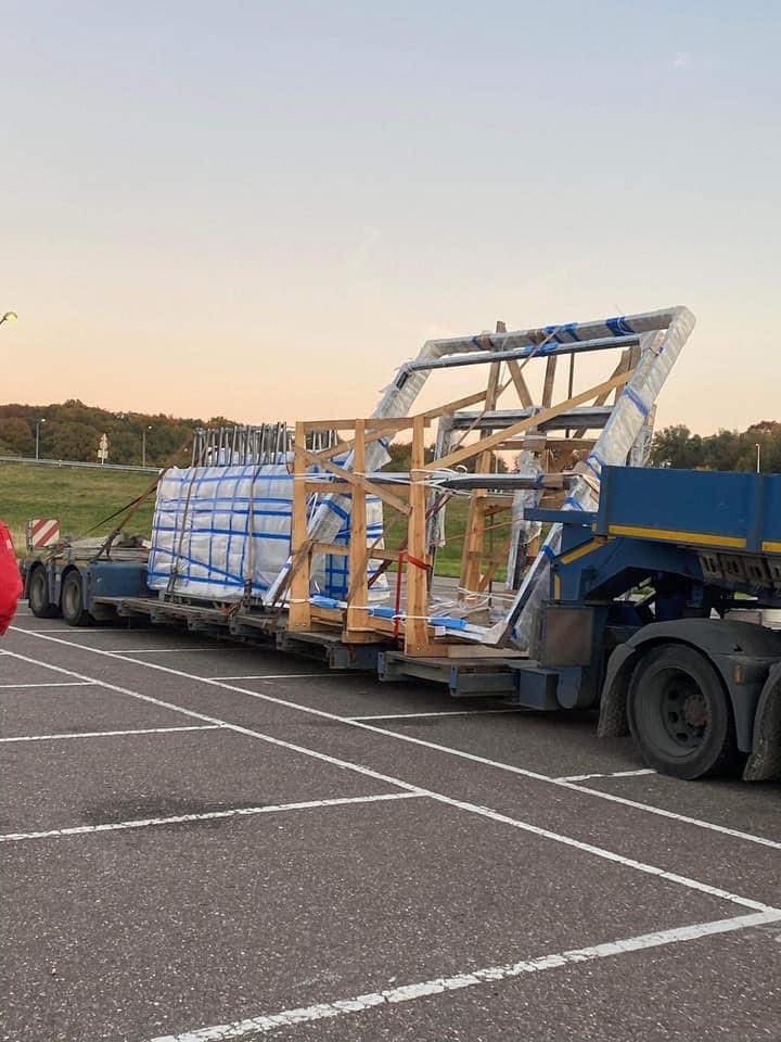 Transport okien w drewnianej ramie na naczepie ciężarówki, zabezpieczone folią, na parkingu z widokiem na zieleń w tle, wczesny poranek.
