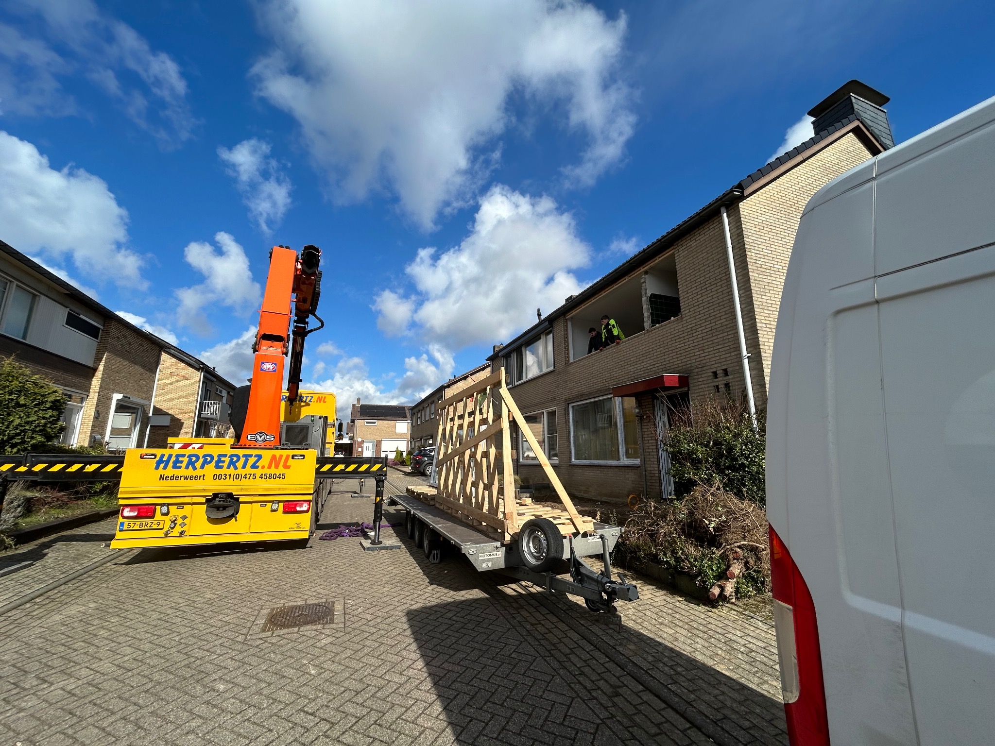 Żuraw Herpertz.nl podnosi drewnianą ramę okienną na przyczepie, w tle budynek z otworem okiennym i pracownicy. Błękitne niebo z chmurami.