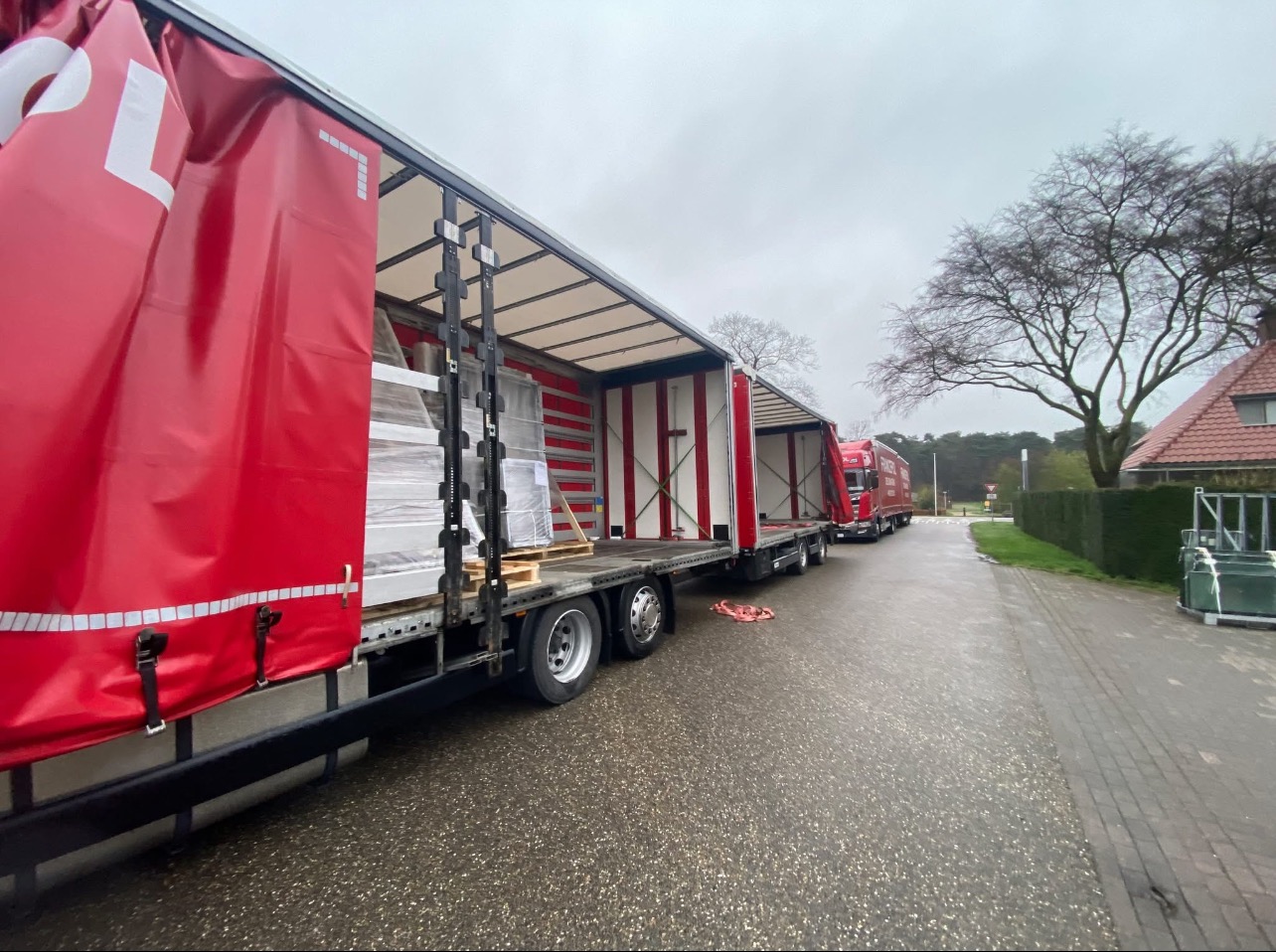 Ciężarówki z naczepami, załadowane oknami i drzwiami, stojące na mokrej drodze w pochmurny dzień. Widoczne detale zabezpieczeń ładunku.