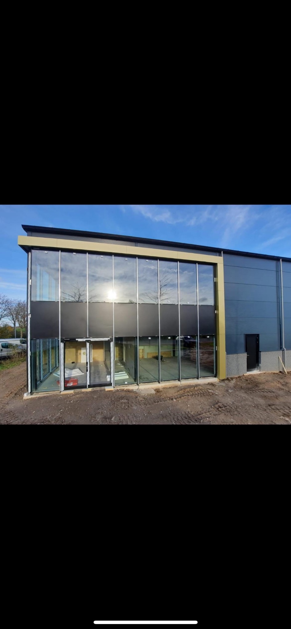 Nowoczesny budynek z fasadą ze szkła i czarnych paneli, odbijający niebo. Widoczne drzwi wejściowe i teren wokół budynku w trakcie prac wykończeniowych.