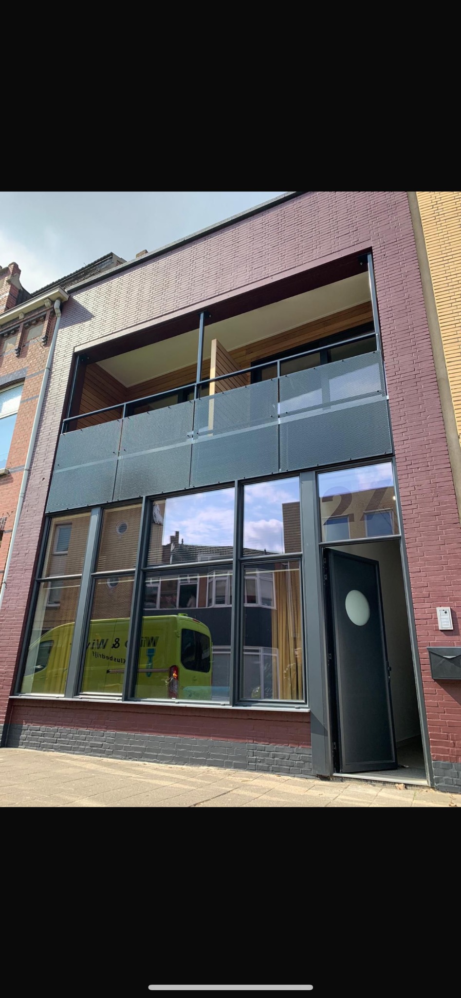 Dwupiętrowy budynek z dużymi oknami i balkonem. Widoczny otwarty czarny drzwi wejściowe z okrągłym oknem. W oknach odbija się zielony samochód dostawczy.