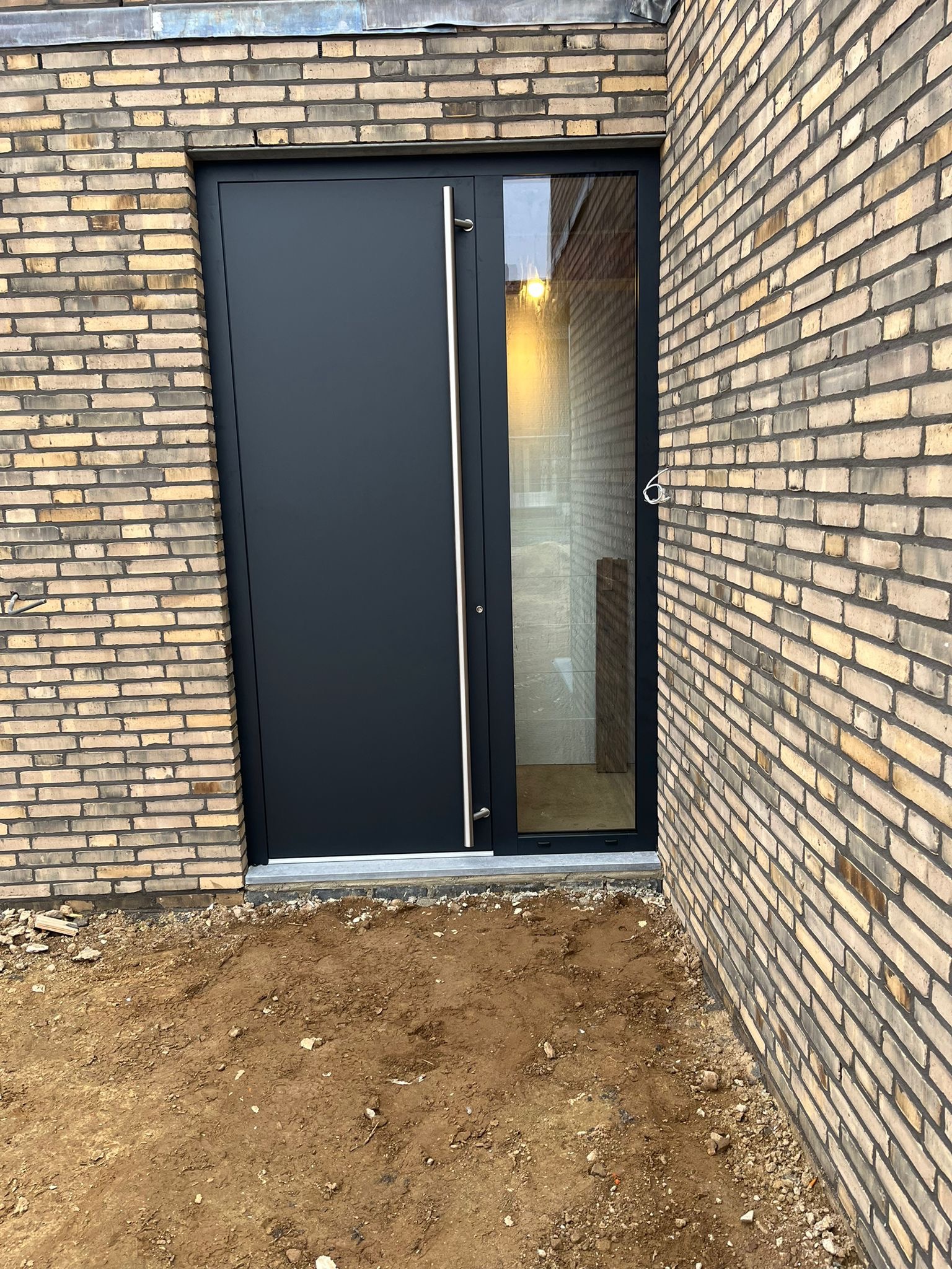 Nowoczesne, czarne drzwi wejściowe z długim, srebrnym uchwytem, wkomponowane w elewację z cegły klinkierowej. Częściowo przeszklone skrzydło.