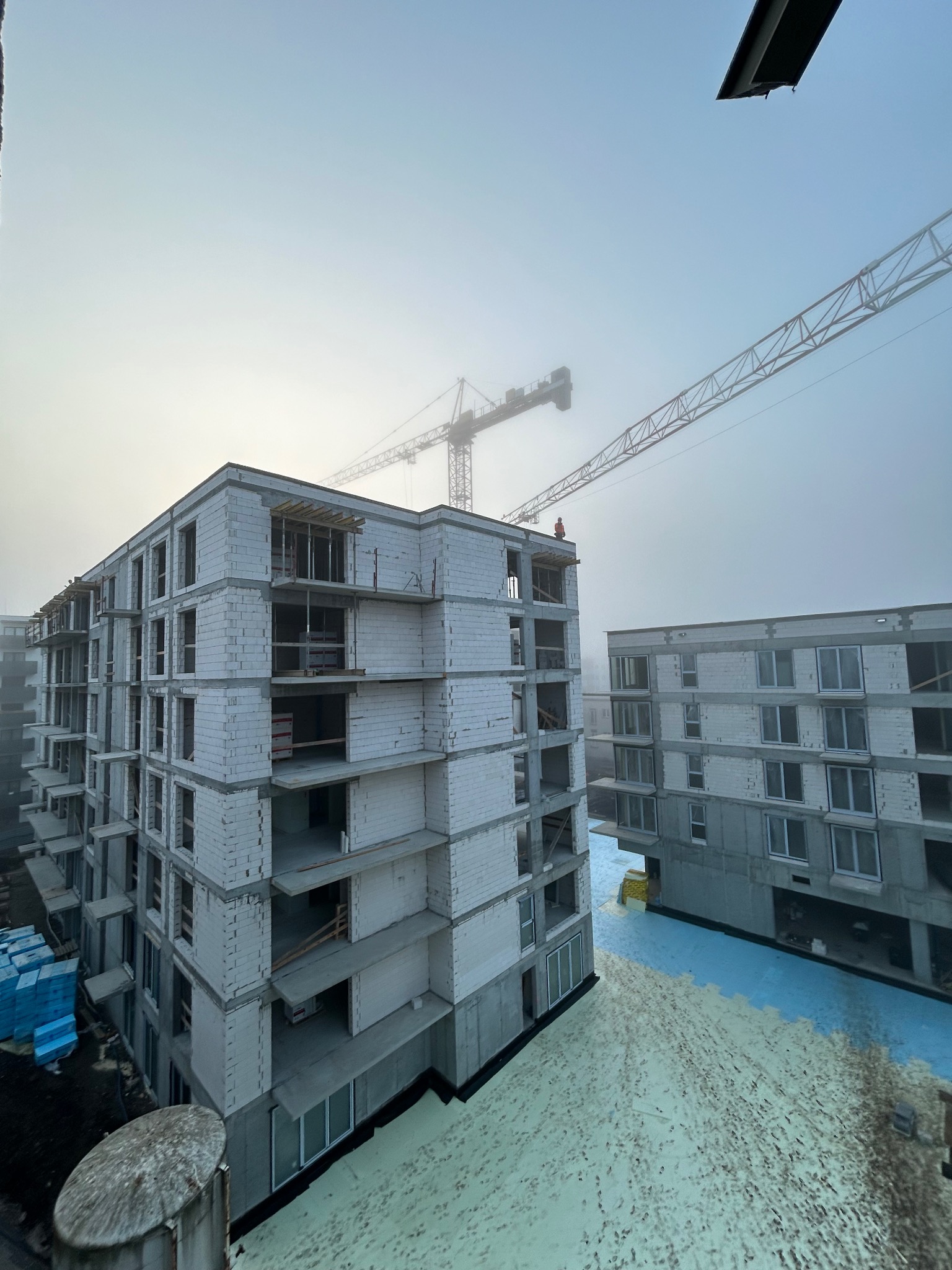 Budowa bloku mieszkalnego w mglisty dzień, widoczne surowe ściany z otworami okiennymi, dźwig budowlany w tle, niebieskie materiały izolacyjne na ziemi.