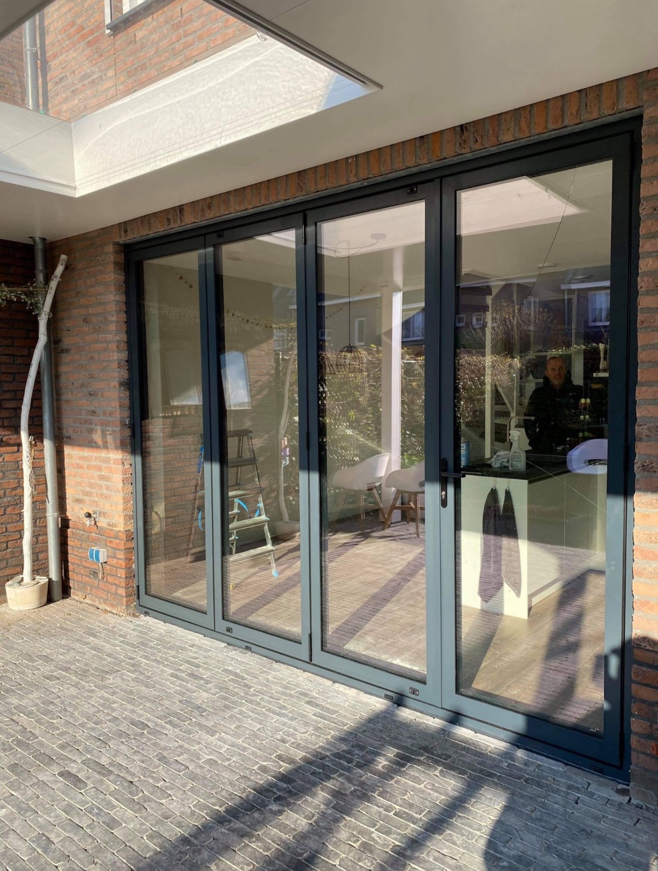 Szklane drzwi przesuwne harmonijkowe z szarymi ramami, widoczne z zewnątrz na tle ceglanego budynku w Czarnkowie. Wnętrze domu widoczne przez szyby.