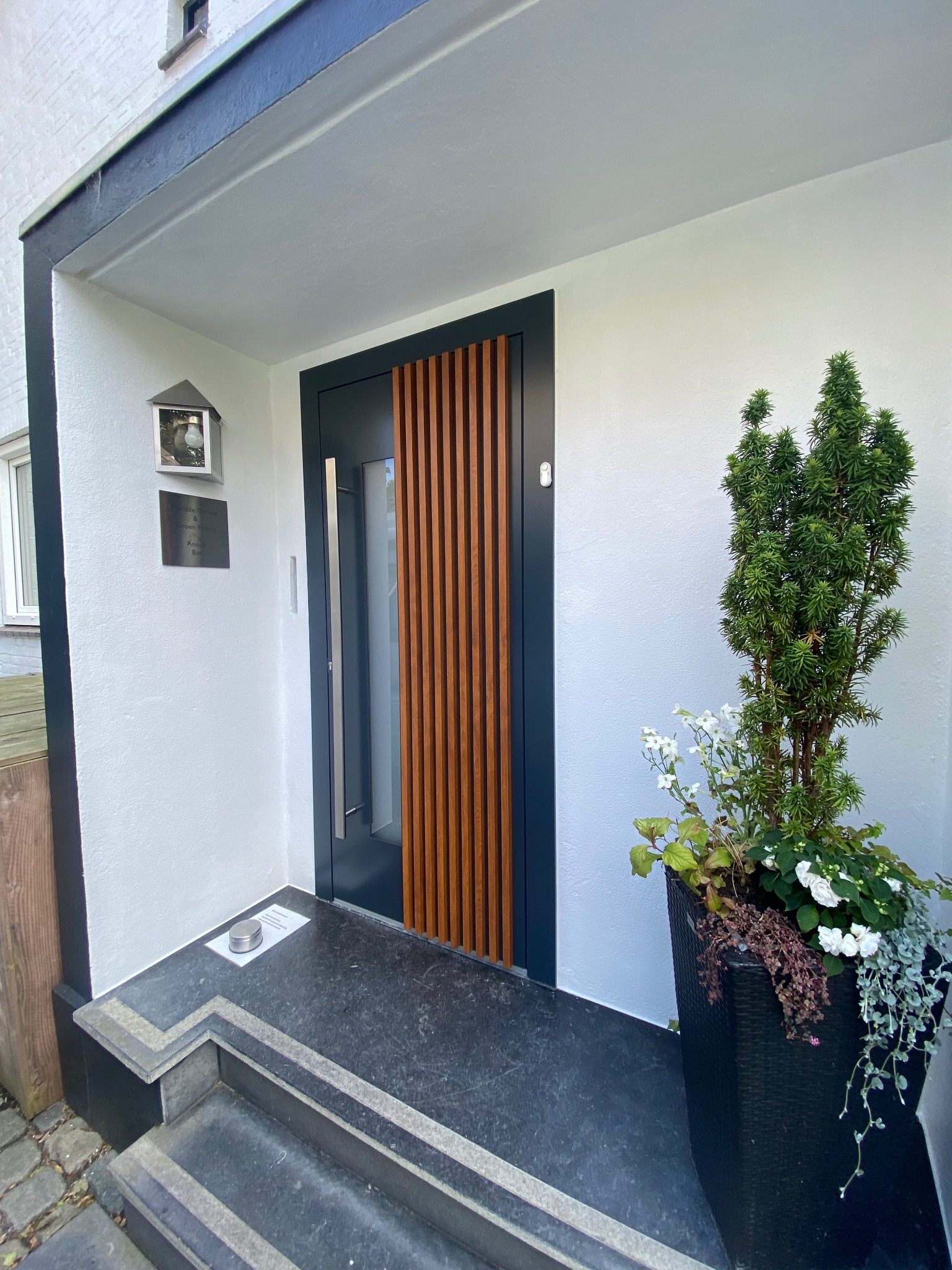 Nowoczesne drzwi wejściowe z drewnianymi lamelami, szara rama, roślina w donicy obok. Wejście do domu z oświetleniem zewnętrznym i tabliczką informacyjną.