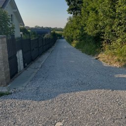 Jan Bednarczyk - Droga szutrowa prowadząca wzdłuż ogrodzenia i zieleni, z widocznym domem w tle. Świeżo wysypany żwir tworzy równą nawierzchnię. Perspektywa z dołu, słoneczny dzień.