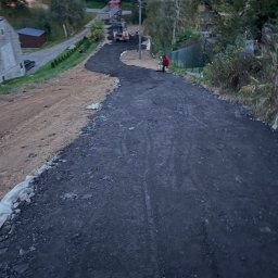 Jan Bednarczyk - Układanie nawierzchni asfaltowej na drodze dojazdowej, widoczny walec i ciężarówka w tle. Częściowo wyrównany teren po prawej stronie.