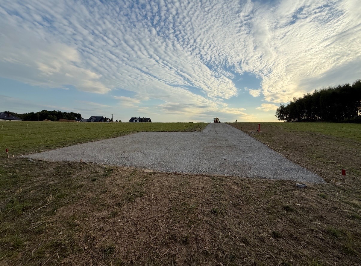 Utwardzony teren żwirowy na działce budowlanej, w tle domy i błękitne niebo z chmurami. Na horyzoncie widoczna maszyna budowlana.