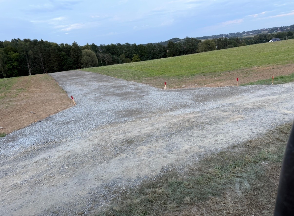 Ułożona warstwa tłucznia na gruncie, wyznaczona palikami. Przygotowanie podłoża pod drogę na nierównym terenie, widok z góry.