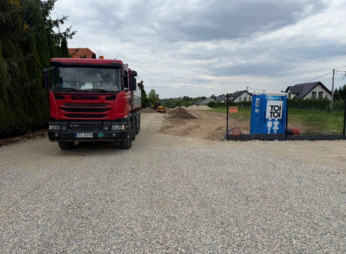 Czerwona ciężarówka Scania na żwirowej drodze w trakcie budowy, w tle TOI TOI i wykop pod drogę. Widoczne domy w oddali.