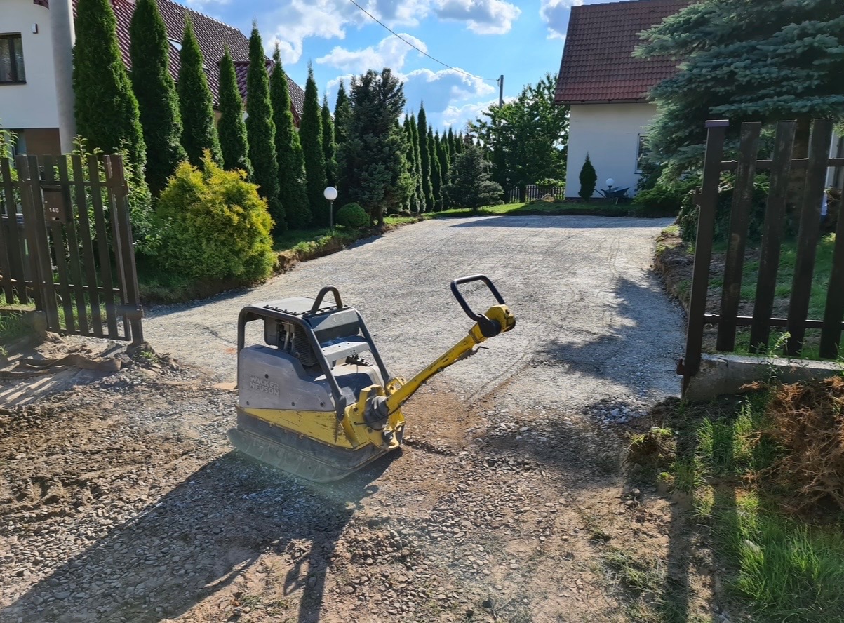 Żółta zagęszczarka spalinowa na żwirowej drodze wjazdowej, częściowo utwardzonej, z widocznymi śladami pracy, otoczonej zielenią i ogrodzeniem.