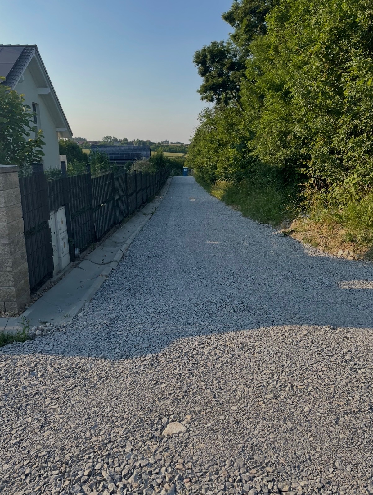 Droga szutrowa prowadząca wzdłuż ogrodzenia i zieleni, z widocznym domem w tle. Świeżo wysypany żwir tworzy równą nawierzchnię. Perspektywa z dołu, słoneczny dzień.