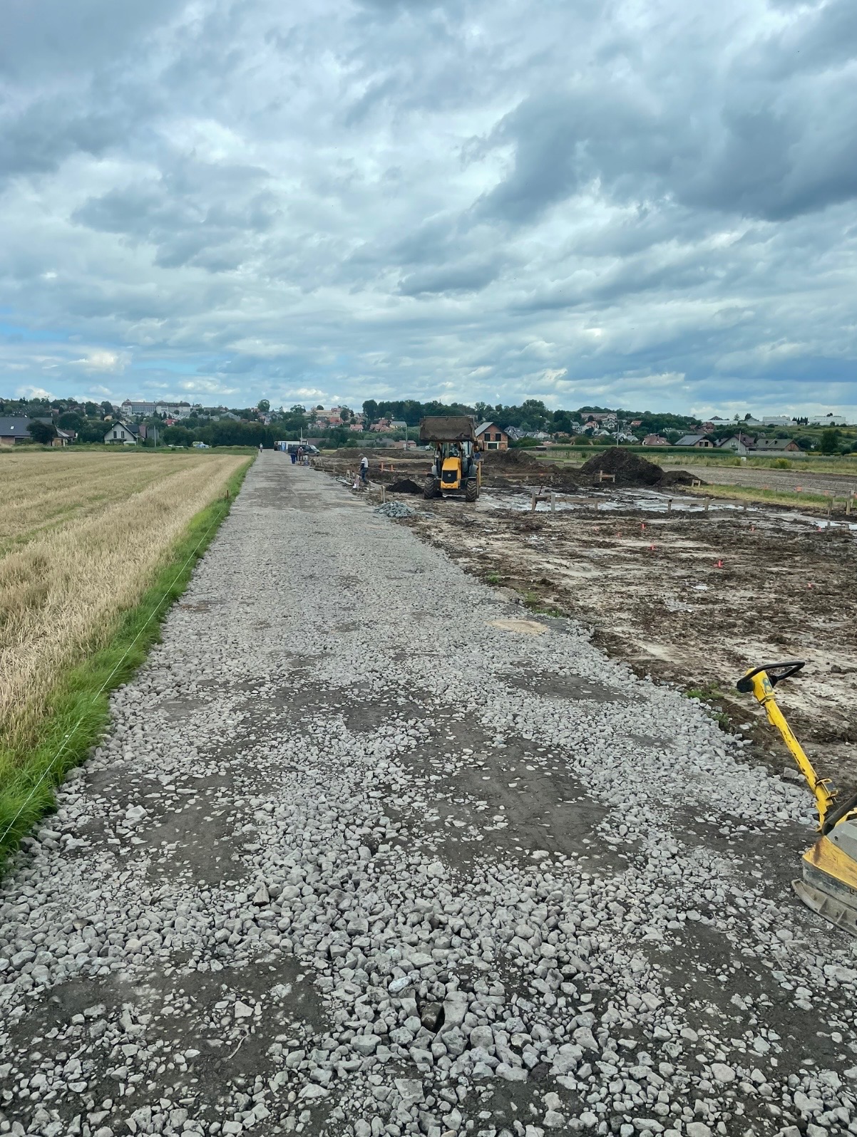 Budowa drogi gruntowej z użyciem walca i koparki na tle pola i zabudowań. Widok na ubitą nawierzchnię z kamieni. Pochmurne niebo.