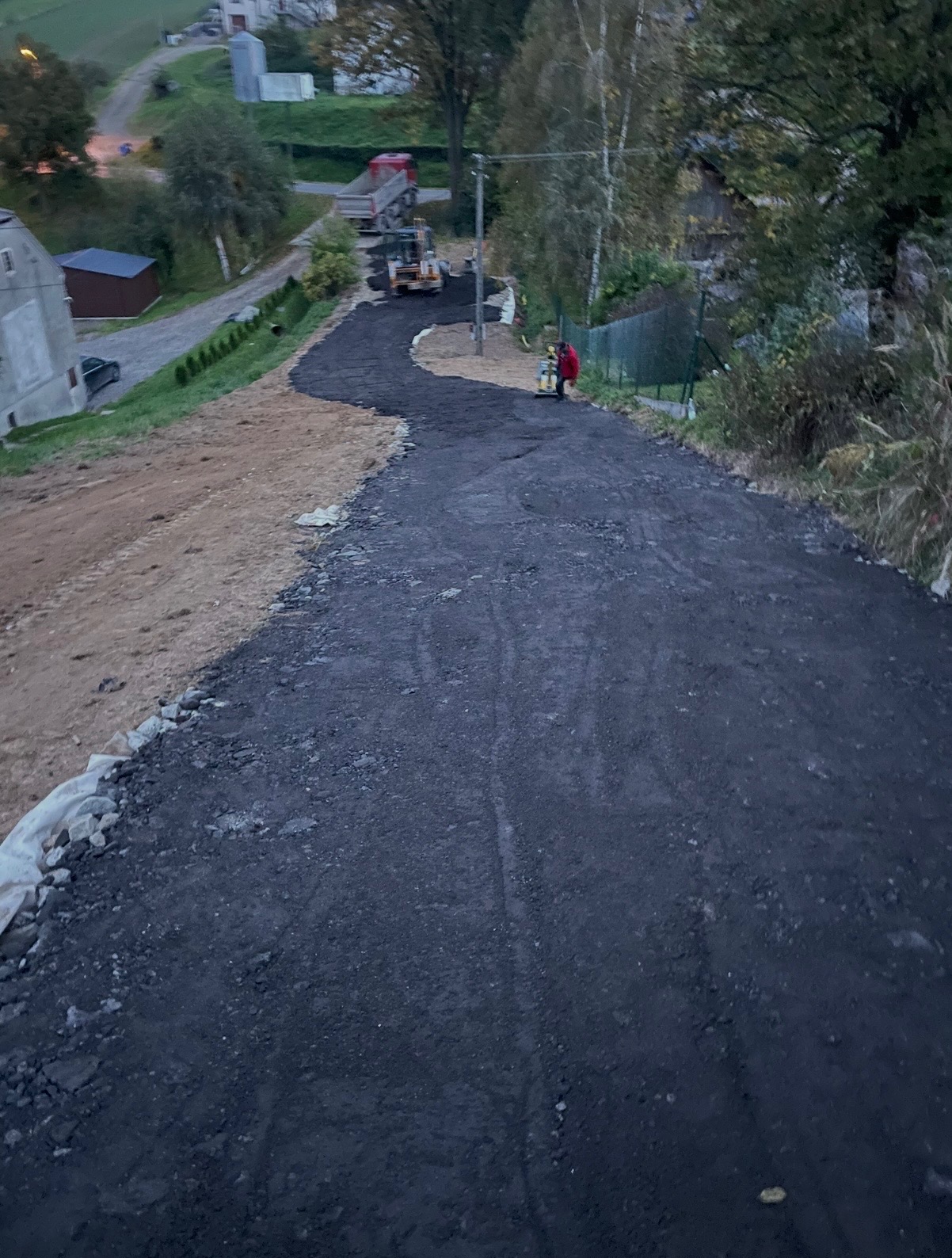 Układanie nawierzchni asfaltowej na drodze dojazdowej, widoczny walec i ciężarówka w tle. Częściowo wyrównany teren po prawej stronie.