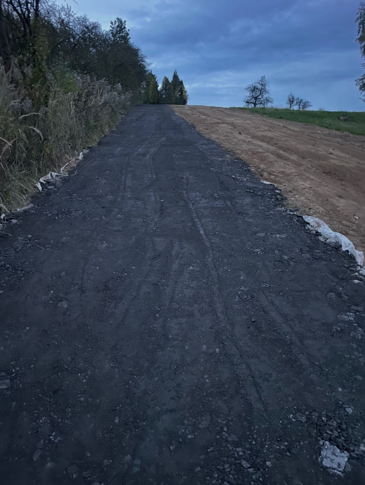 Świeżo położona, czarna nawierzchnia drogi gruntowej, kontrastująca z jasną ziemią po prawej stronie. Widoczne ślady opon i roślinność wzdłuż krawędzi.