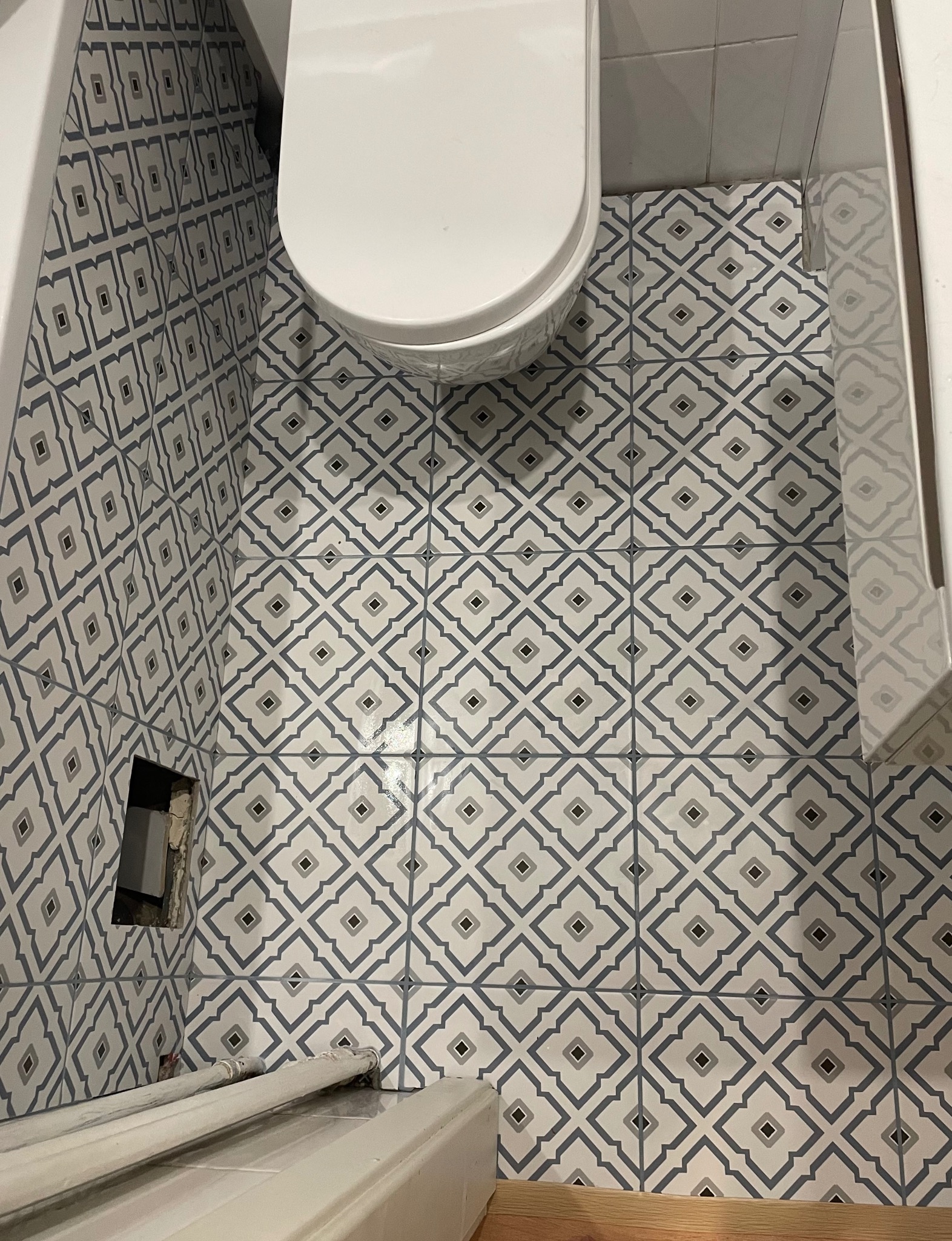 Wzorzyste, szaro-białe płytki w łazience, ułożone na podłodze i ścianach. Widoczny fragment toalety i niedokończone wykończenie ściany z otworem. Ujęcie z góry.