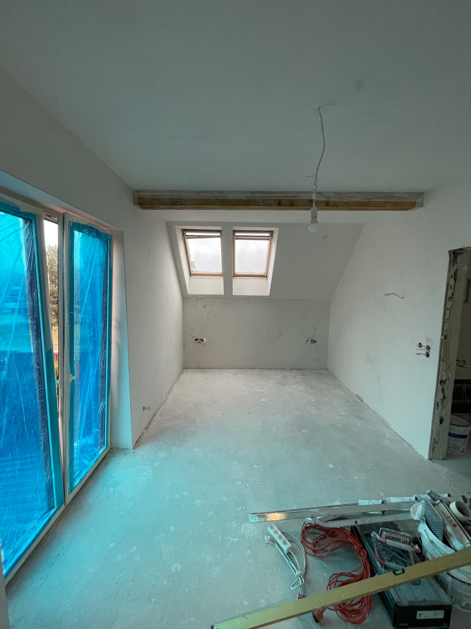 Pokój na poddaszu w trakcie prac wykończeniowych: okna zabezpieczone folią, ściany przygotowane do malowania, widoczne narzędzia i kable na podłodze. Dwa okna dachowe.