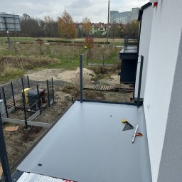 Sokół Budownictwo - Hydroizolacja tarasu membramą PVC na płytach PIR. Widoczne narzędzia: miarka, kątownik, nożyczki. W tle budynki mieszkalne i drzewa. Pochmurne niebo.