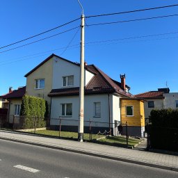 Sokół Budownictwo - Elewacja domu po malowaniu, z nową werandą w surowym drewnie i betonowymi schodami wejściowymi. Widok z ulicy, błękitne niebo w tle.