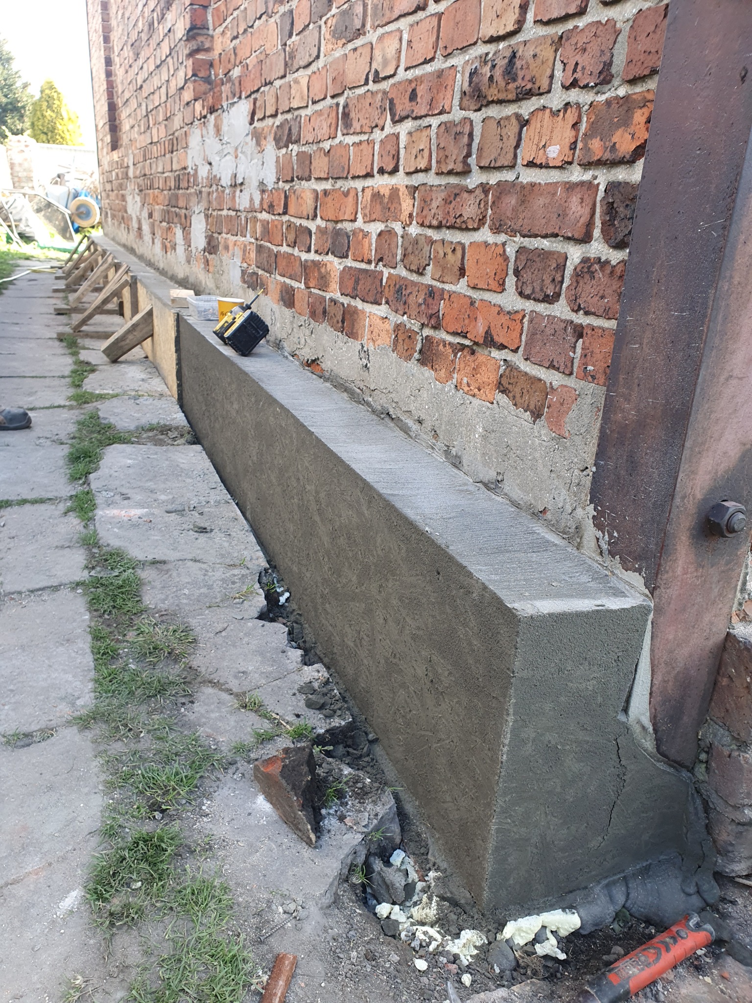 Wzmocniony fundament budynku z opaską zbrojonego betonu, sięgającą ponad grunt. Widoczna ceglana ściana budynku i narzędzia budowlane.