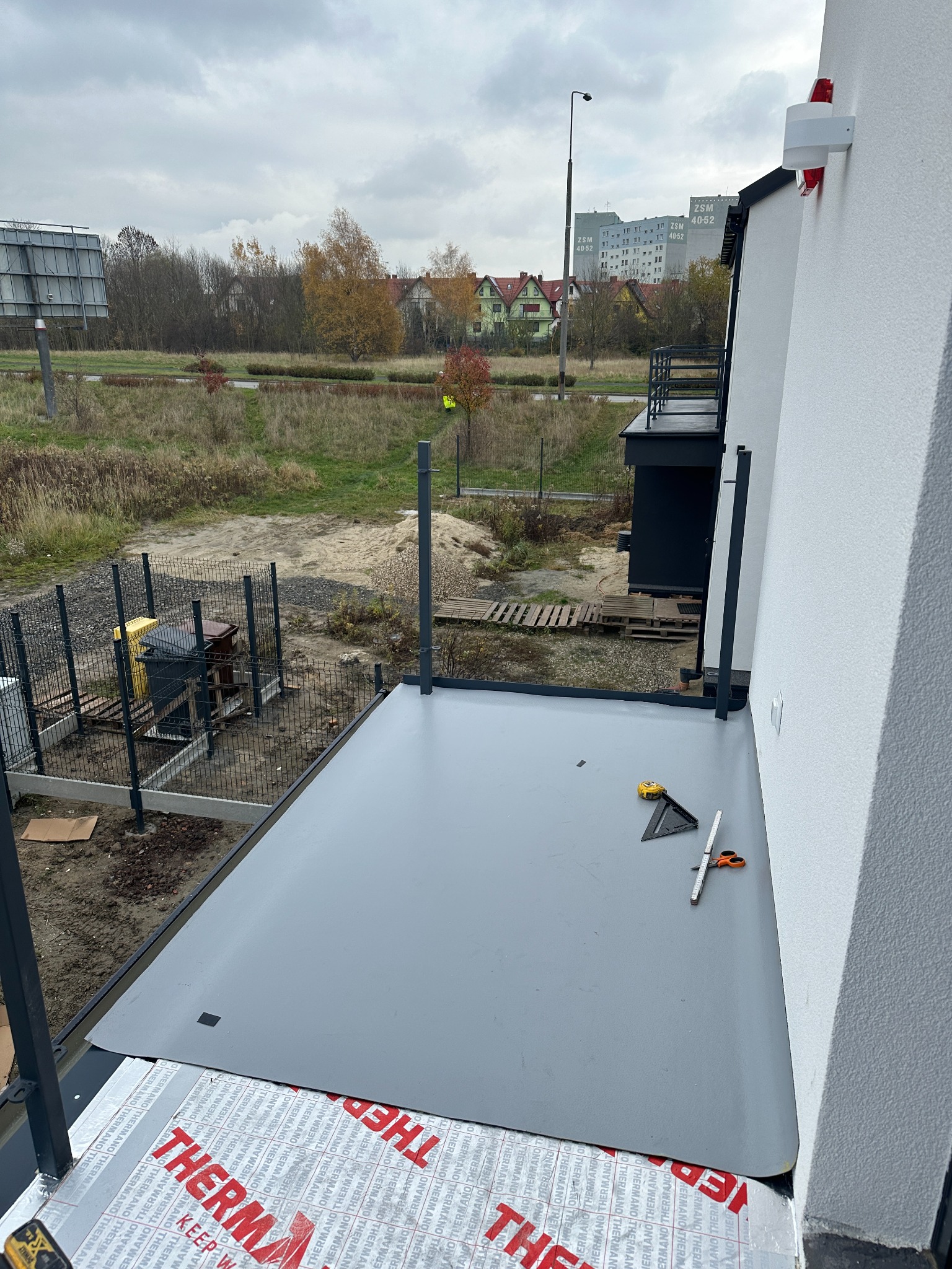 Hydroizolacja tarasu membramą PVC na płytach PIR. Widoczne narzędzia: miarka, kątownik, nożyczki. W tle budynki mieszkalne i drzewa. Pochmurne niebo.