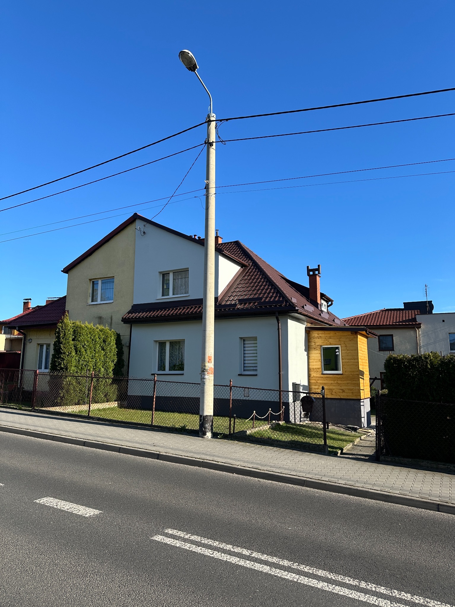 Elewacja domu po malowaniu, z nową werandą w surowym drewnie i betonowymi schodami wejściowymi. Widok z ulicy, błękitne niebo w tle.