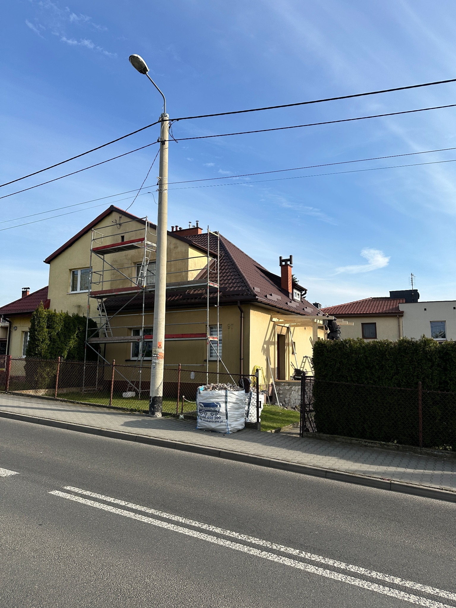 Żółty dom jednorodzinny w trakcie remontu elewacji, z widocznymi rusztowaniami i workiem materiałów budowlanych. Czerwony dach i błękitne niebo w tle.