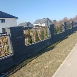 Marlena Okapa - Nowoczesne ogrodzenie z szarej kostki i metaloplastyki, oddzielające posesję z tujami od chodnika. W tle domy jednorodzinne w słoneczny dzień.