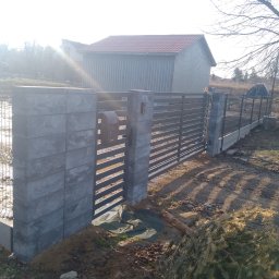 Marlena Okapa - Nowoczesne ogrodzenie panelowe z betonowymi słupkami i furtką, skrzynka na listy. W tle budynek gospodarczy i drzewa bez liści, słoneczny dzień.