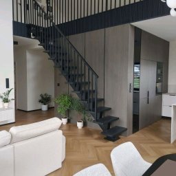 Marlena Okapa - Nowoczesne, minimalistyczne schody z czarną balustradą w jasnym wnętrzu. Widoczne rośliny doniczkowe i parkiet w jodełkę. Uwagę przykuwa nietypowa konstrukcja stopni.