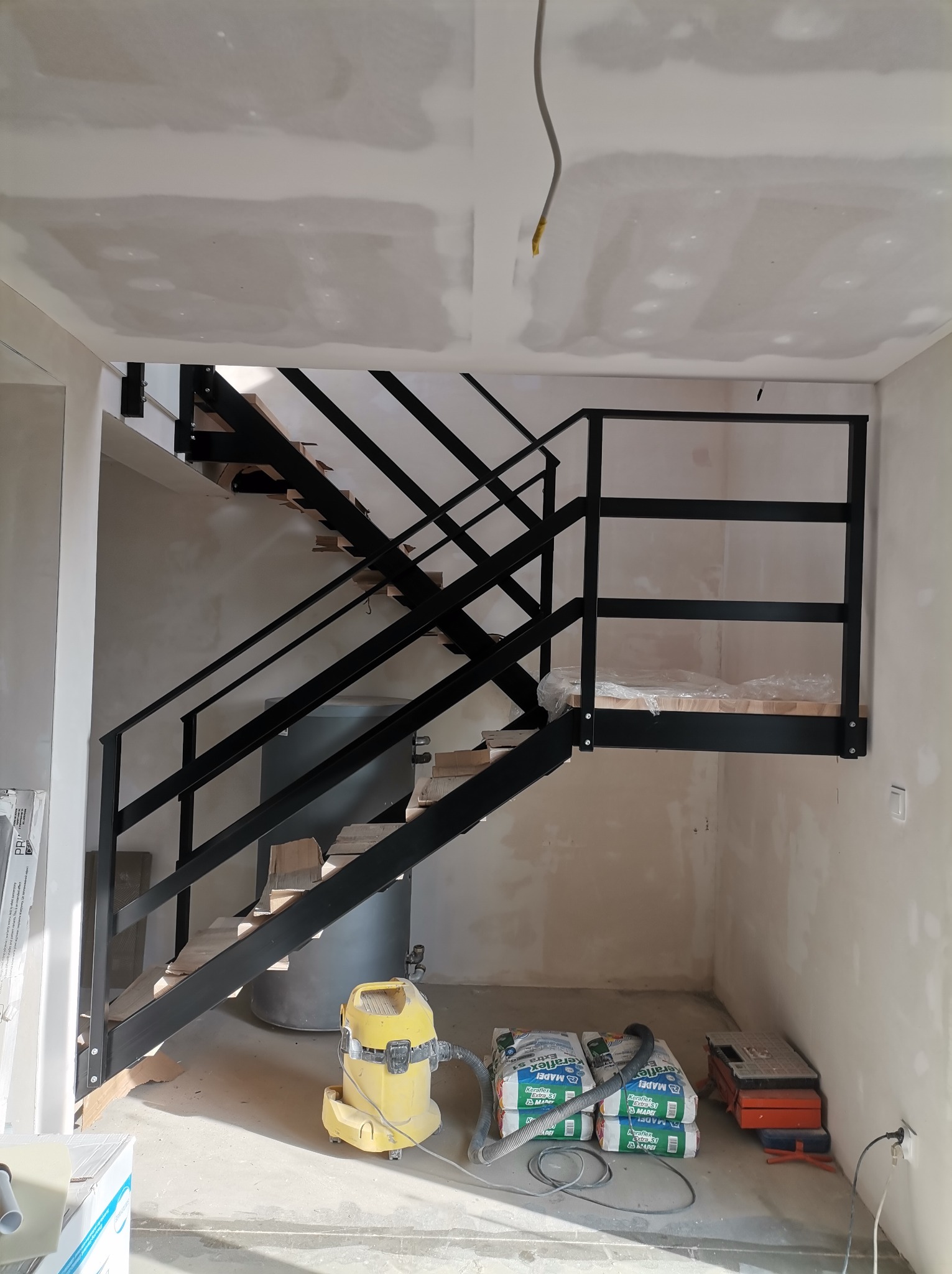 Wnętrze w trakcie wykańczania z zamontowanymi czarnymi schodami z metalową balustradą. Widoczne narzędzia i materiały budowlane. Surowy stan ścian i sufitu.