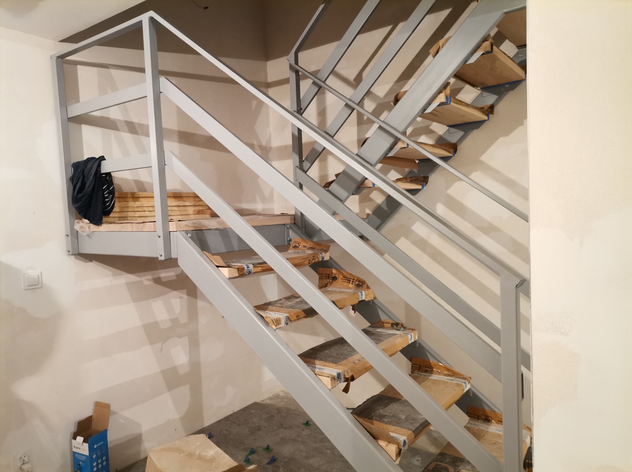 Szare, metalowe schody wewnętrzne z drewnianymi stopniami w trakcie montażu, zabezpieczone kartonem. Widoczne elementy mocujące i narzędzia.