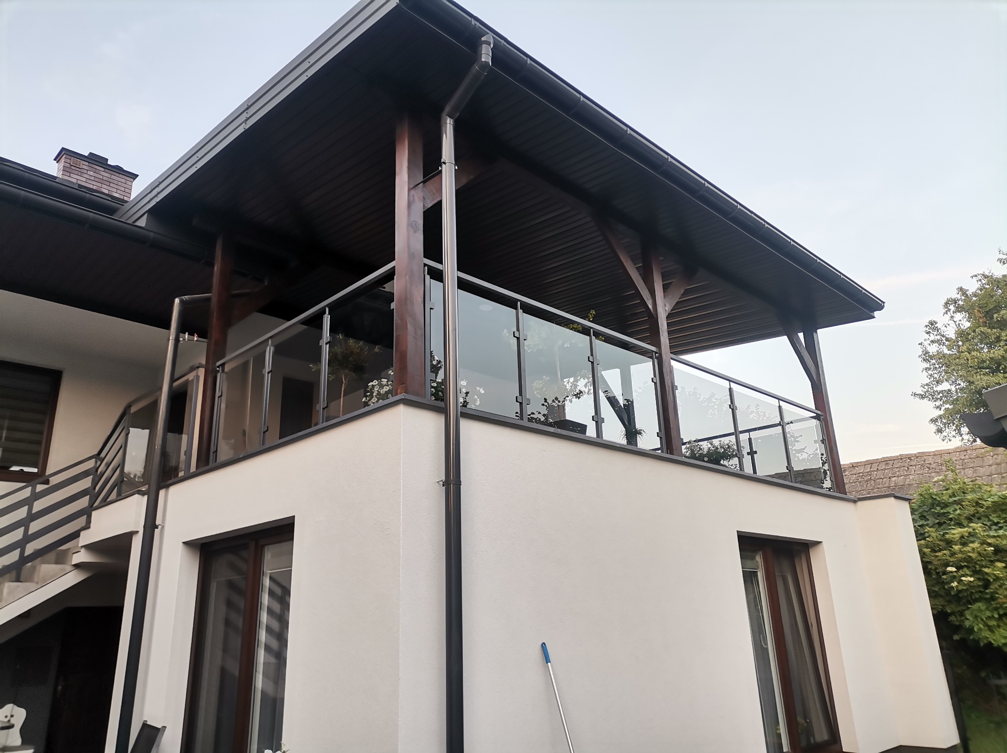 Nowoczesny balkon z drewnianą konstrukcją i szklanymi balustradami, widok z dołu, jasna elewacja budynku, schody zewnętrzne z metalową barierką.